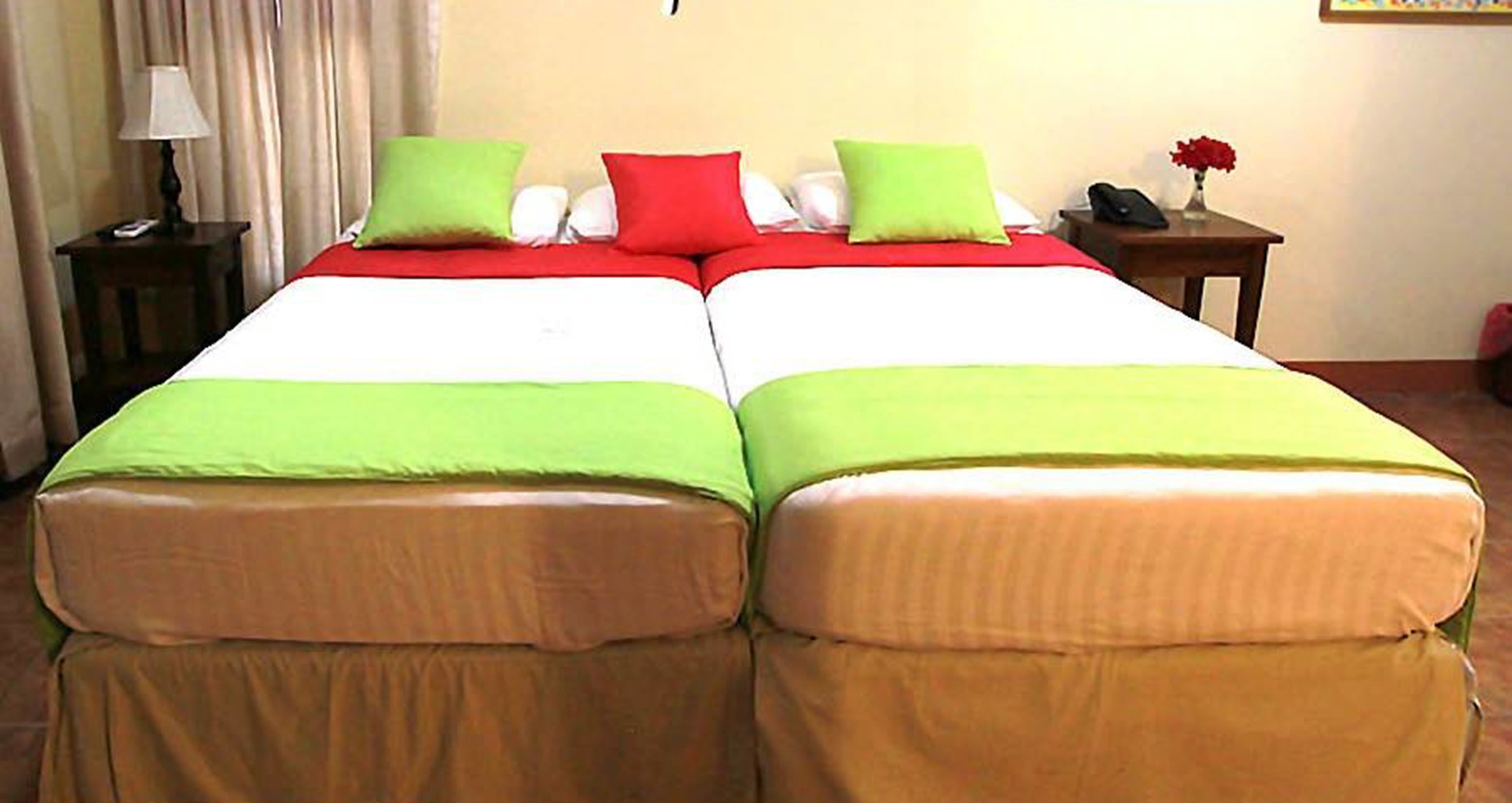 A double bed with green and red bedding, a bedside table with a lamp, and a vase with flowers on the right side.