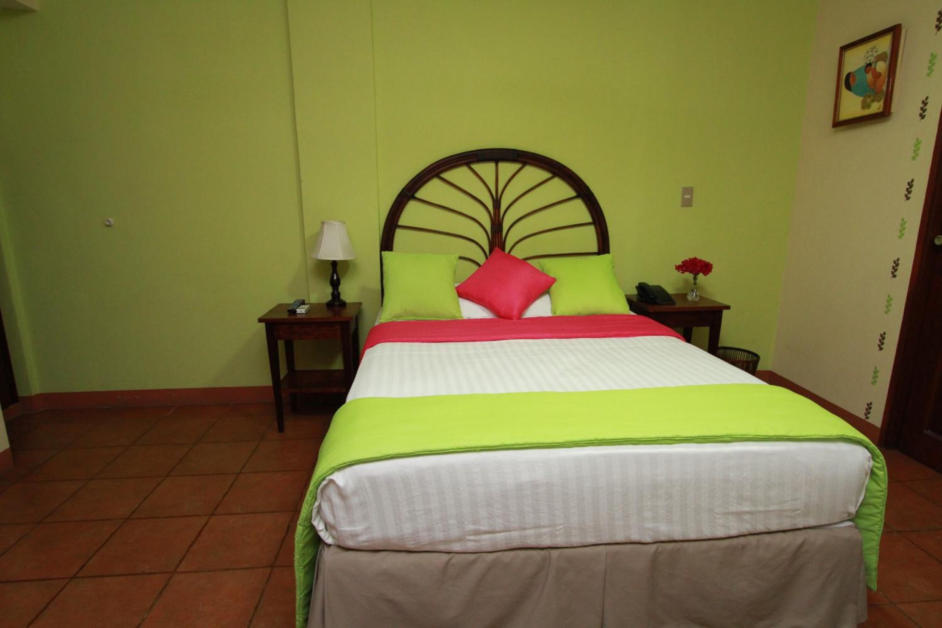 Una cama con colcha verde y blanca, un almohadón rojo y un cabecero de madera. Hay una mesita de noche con una lámpara y un florero.