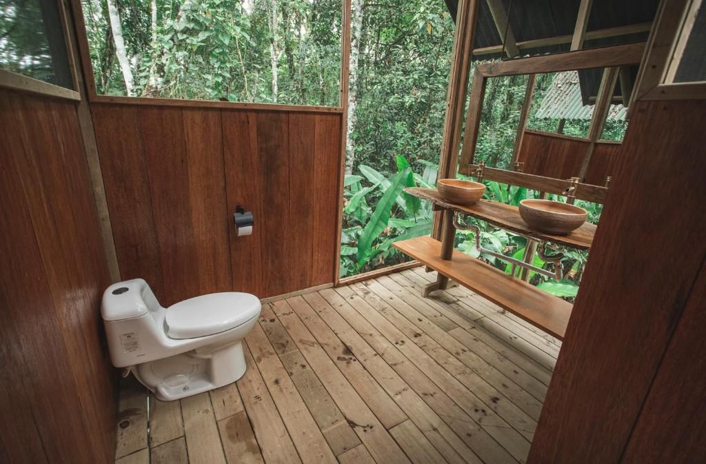 Casa de banho com soalho em madeira, vaso sanitário, prateleiras de madeira e janela com vista para uma área arborizada.