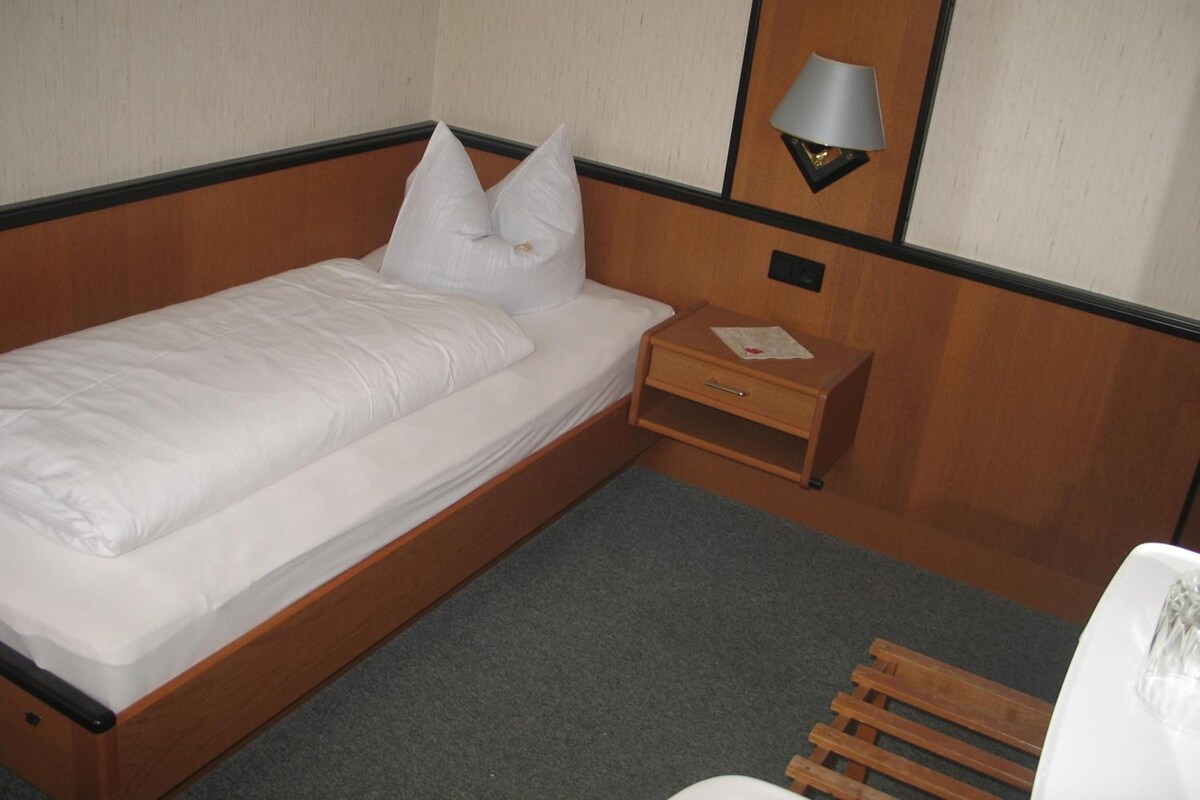 A single bed with a wooden frame, a nightstand with a lamp, and a small drawer.
