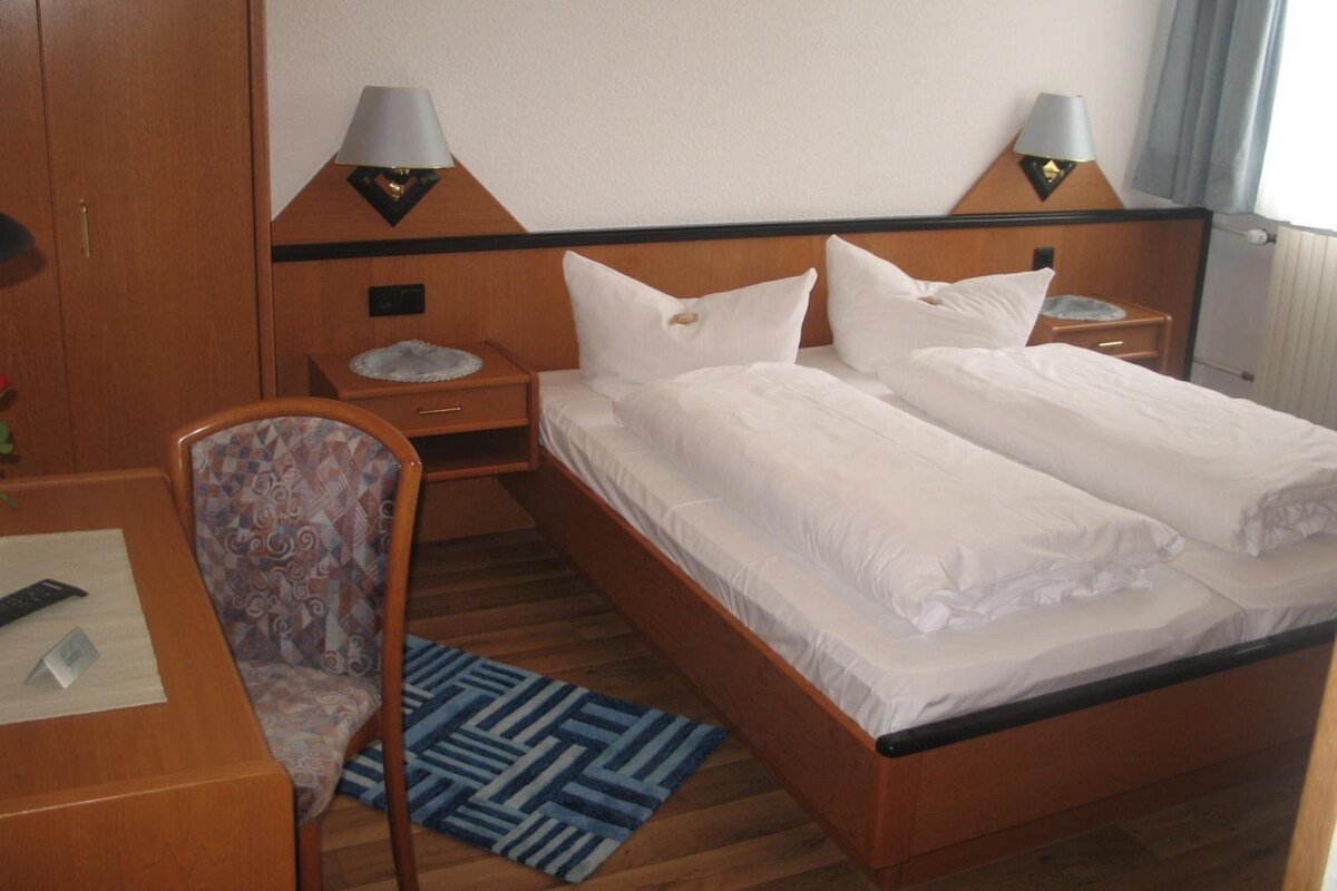 A double bed with white linens, two bedside lamps, a wooden headboard, a small bedside table with a lamp, a patterned chair, and a blue rug.