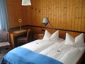 A hotel room with a wooden paneled wall, a bed with white linens, a desk with a chair, and a lamp. - Gasthof Pension Leupold (Selbitz)