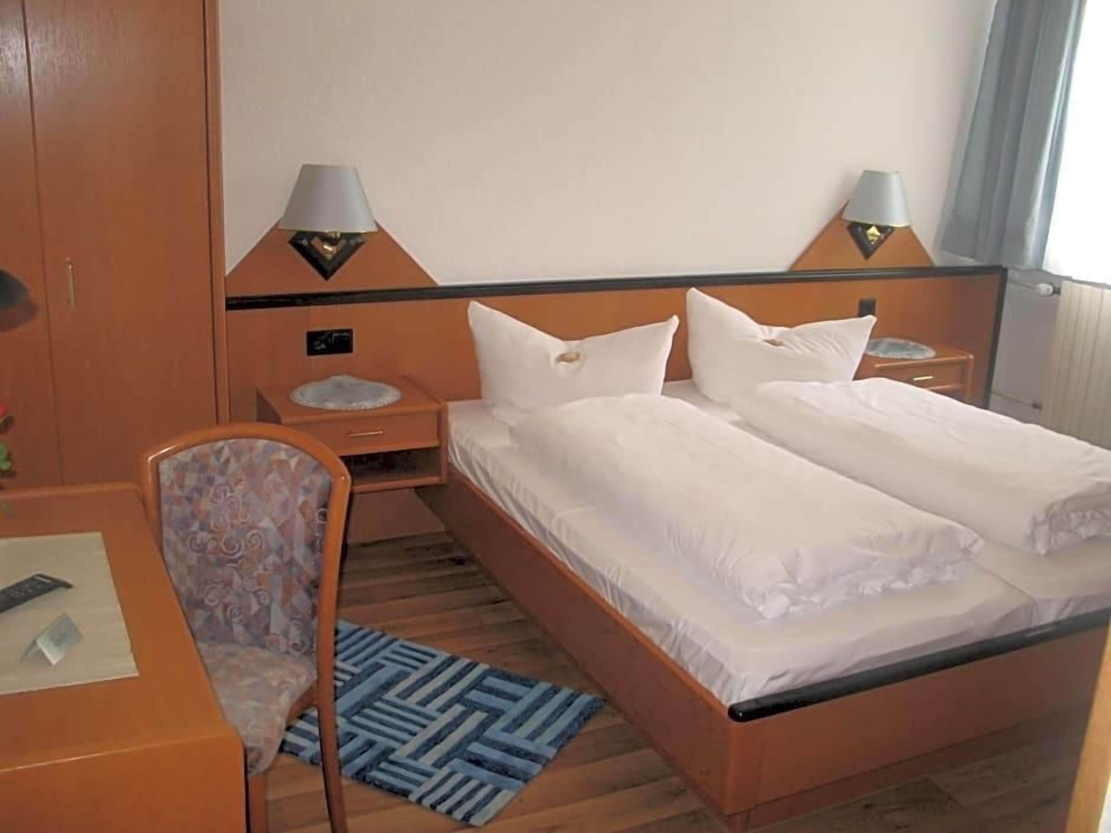 Ein Hotelzimmer mit einem Doppelbett, zwei Nachttischen inklusive Lampen, einem Sessel und einem Schreibtisch mit Fernbedienung.