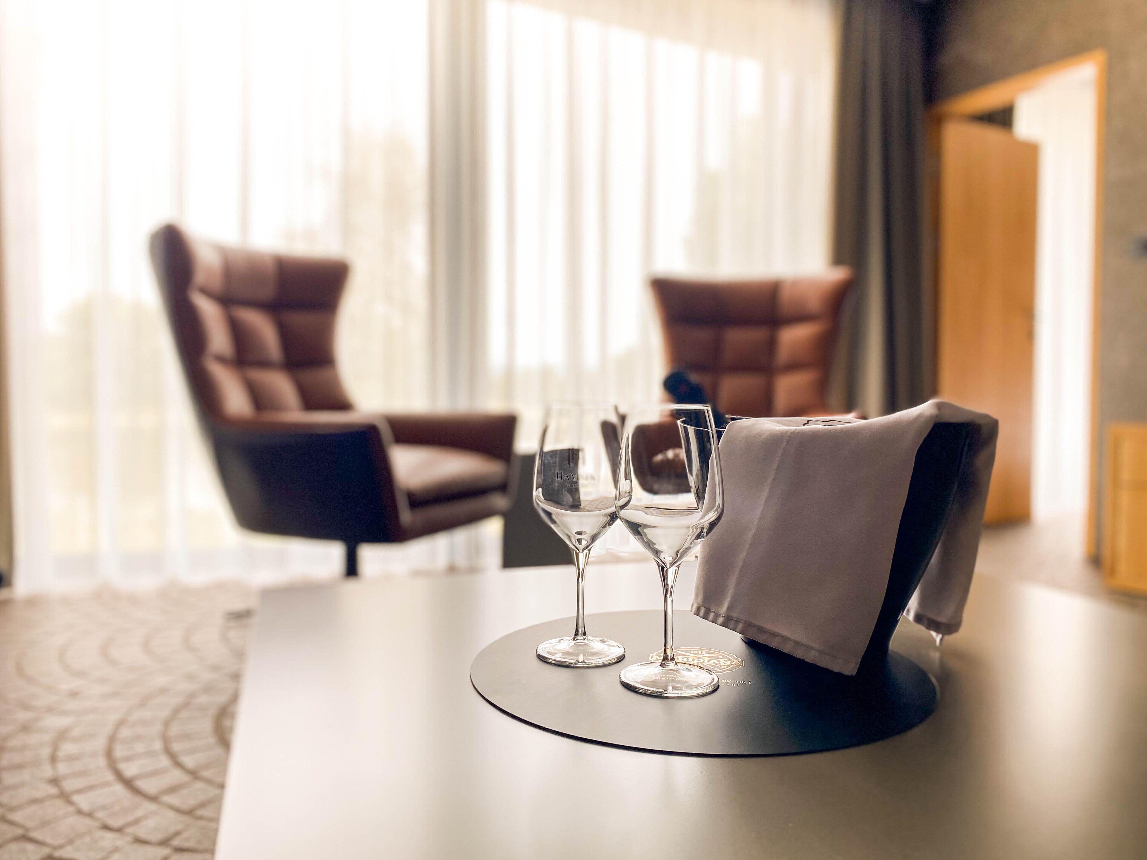 A hotel room with a dining table set for two, two wine glasses, and a folded napkin on a black coaster.