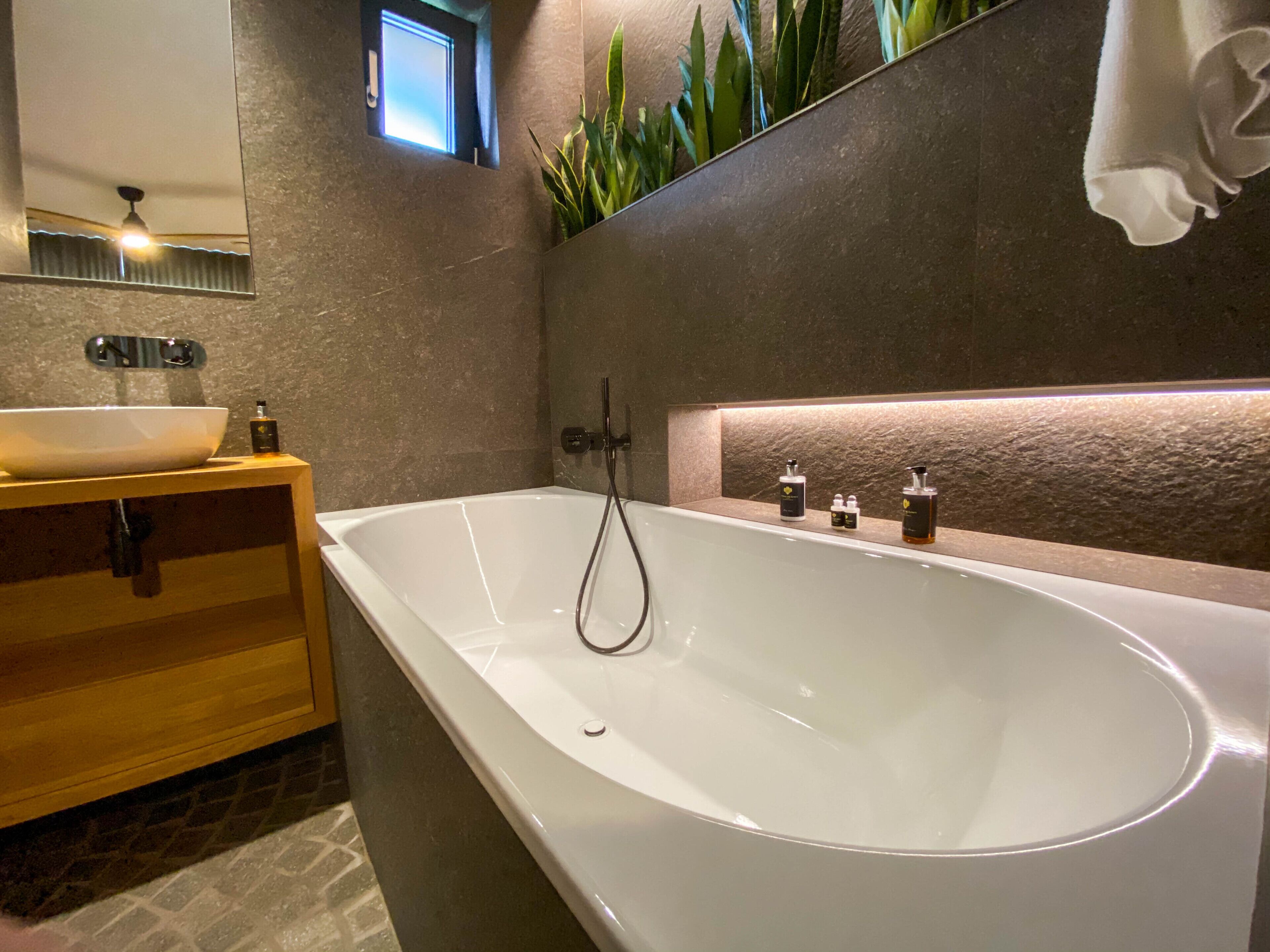 A modern bathroom with a freestanding bathtub, a wooden vanity, a large mirror, and a small window.