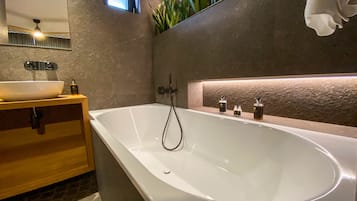 A modern bathroom with a freestanding bathtub, a wooden vanity, a large mirror, and a small window.