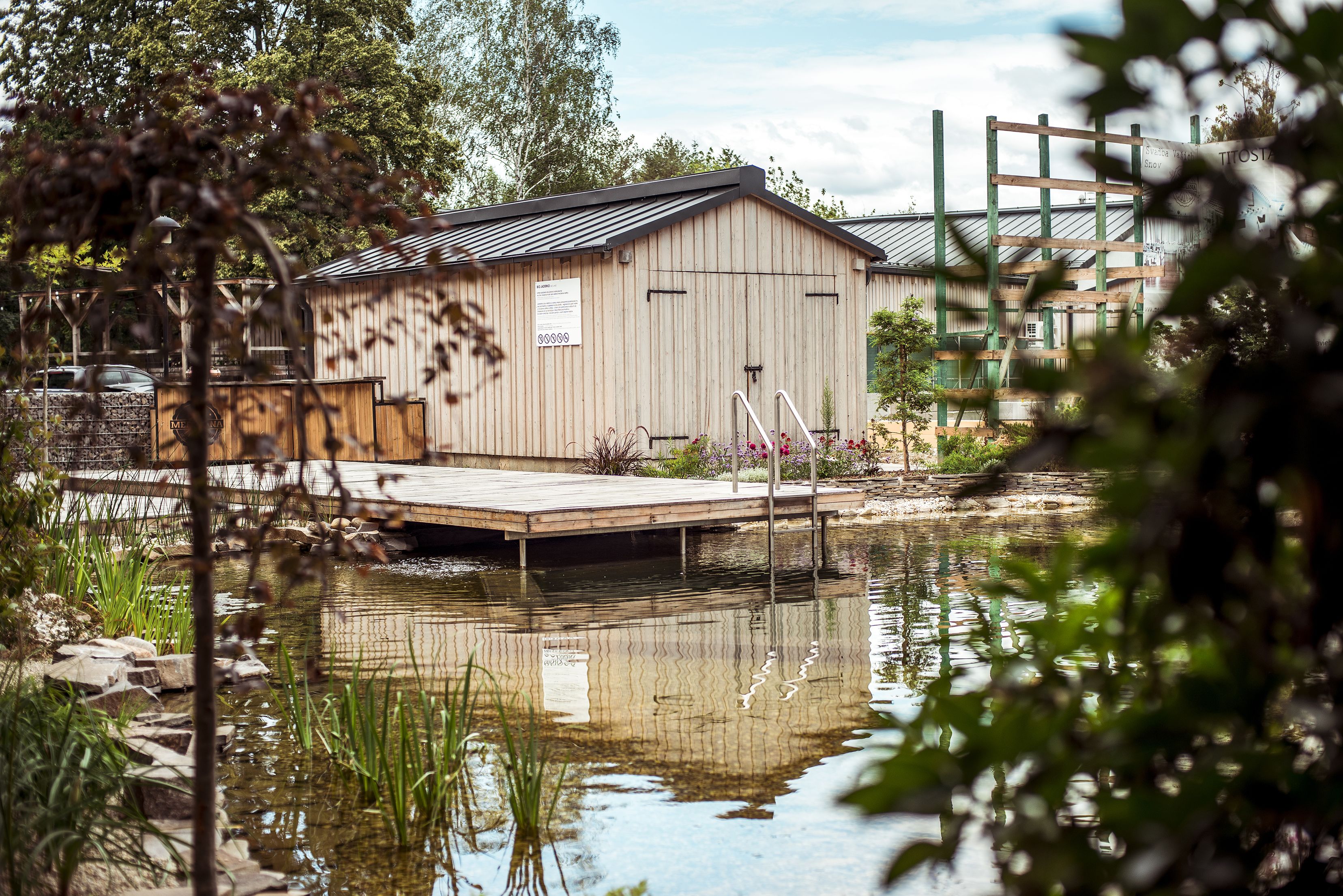 A wooden shed with a dock over a pond, surrounded by greenery and trees.