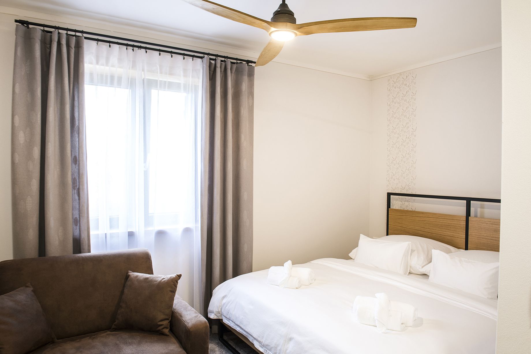 Una habitación de hotel con cama, una silla, ventilador de techo y ventana con cortinas.