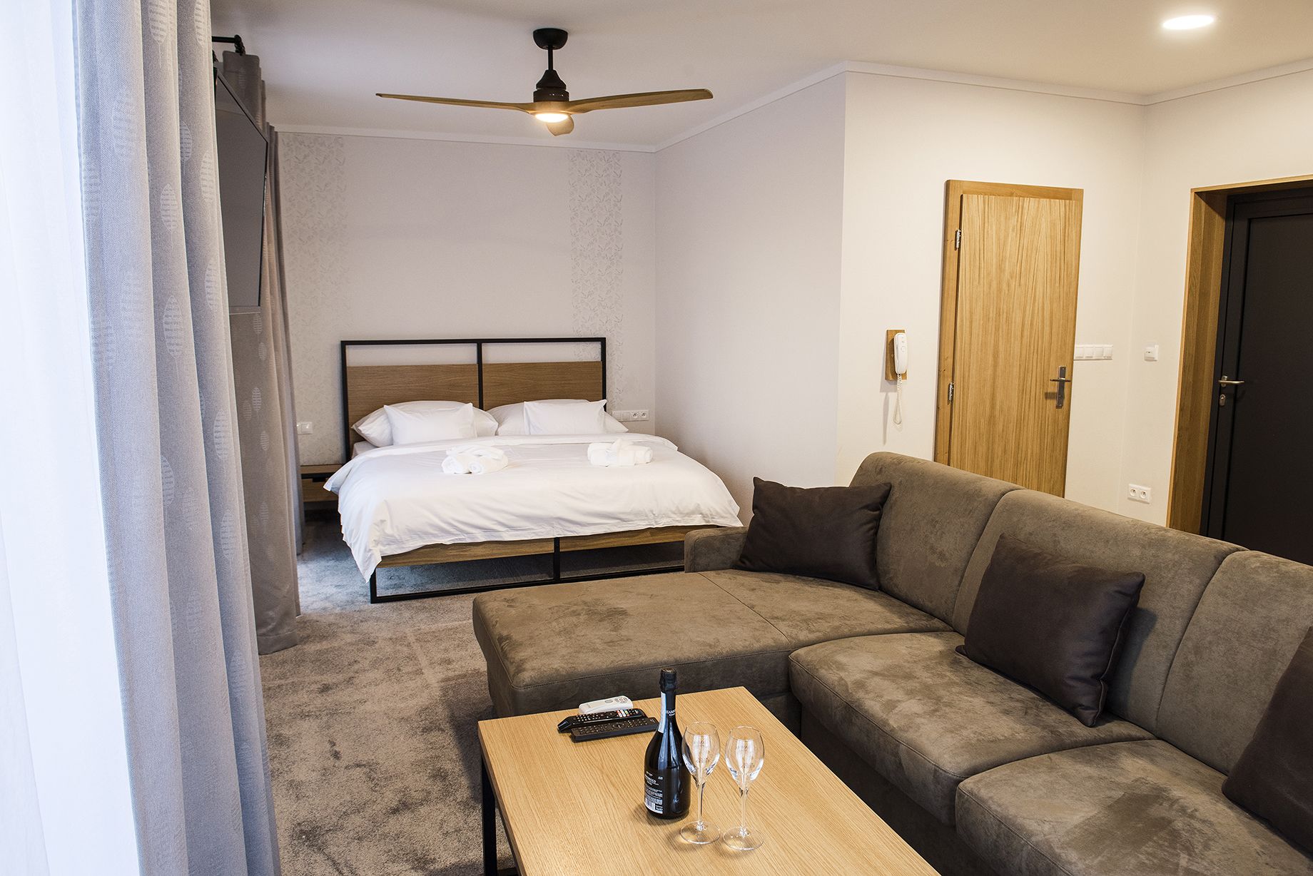 Hotellihuone, jossa on sänky, sohva, pöytä, jonka päällä on pullo ja laseja, sekä kattotuuletin.