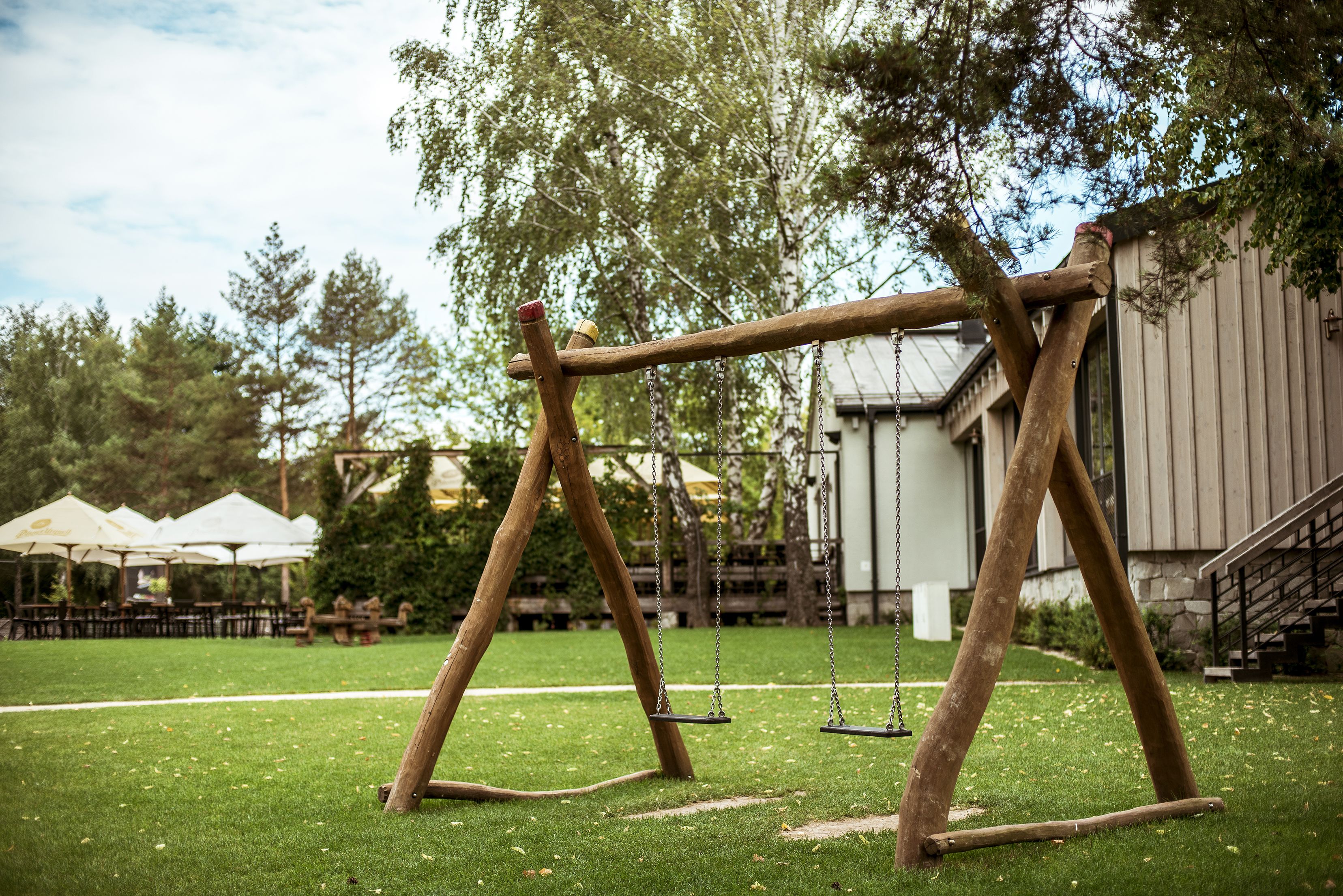 Een houten schommelset in een grasveld, met een gebouw en terrasjes op de achtergrond.