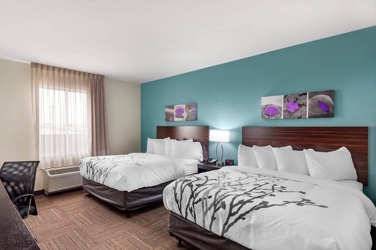 A hotel room with two beds, a desk, and a chair. There are two bedside tables with lamps, a window with curtains, and artwork on the wall.