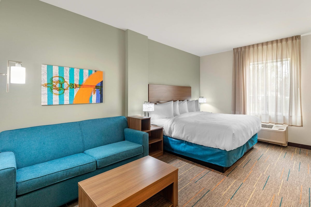 A hotel room with a bed, a blue sofa, a wooden coffee table, a nightstand, a lamp, and a large window with curtains.