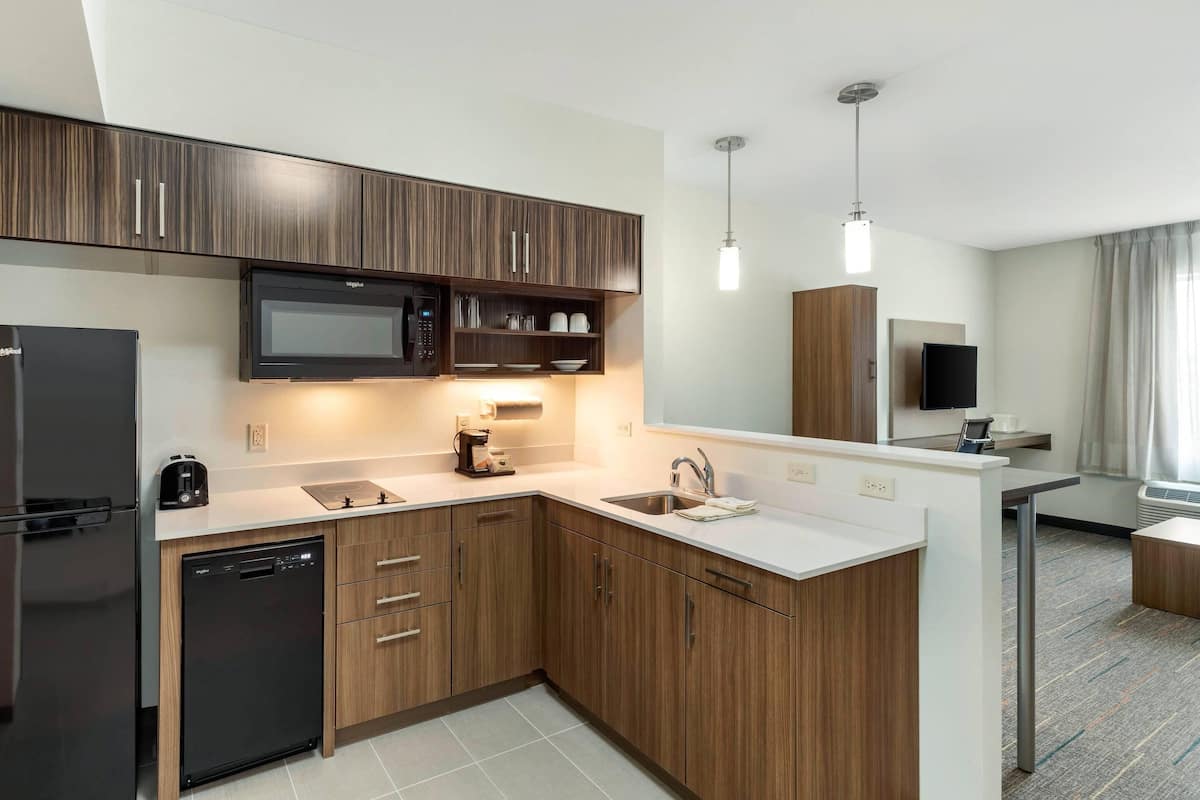 A modern kitchen with wooden cabinets, a microwave, a dishwasher, and a sink. There is a small island with a sink and a flat-screen TV mounted on the wall.