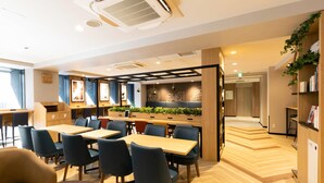 A modern dining area with wooden tables and chairs, a kitchen counter with plants, and a bookshelf. - Comfort Hotel Shin-Osaka (Osaka)