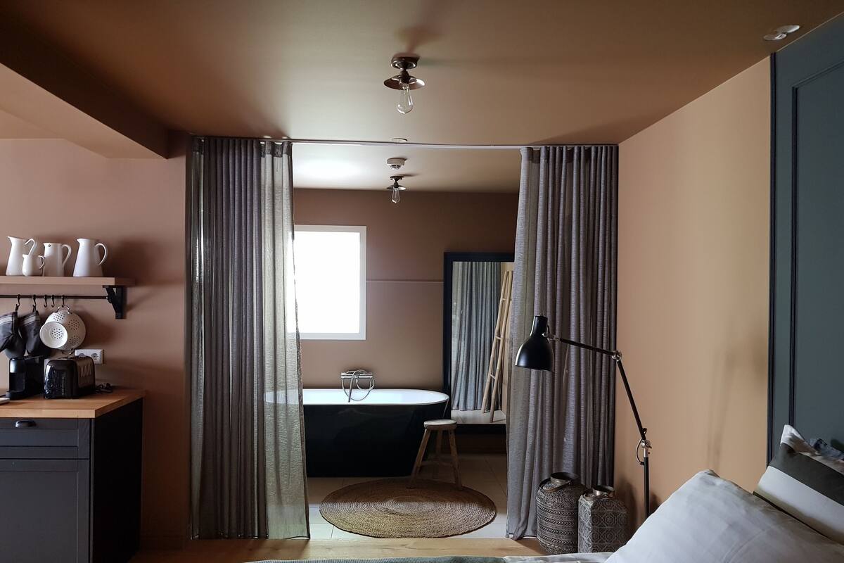 A modern bedroom with a bed, bedside table, lamp, and a view into another room with a dining table and chairs.