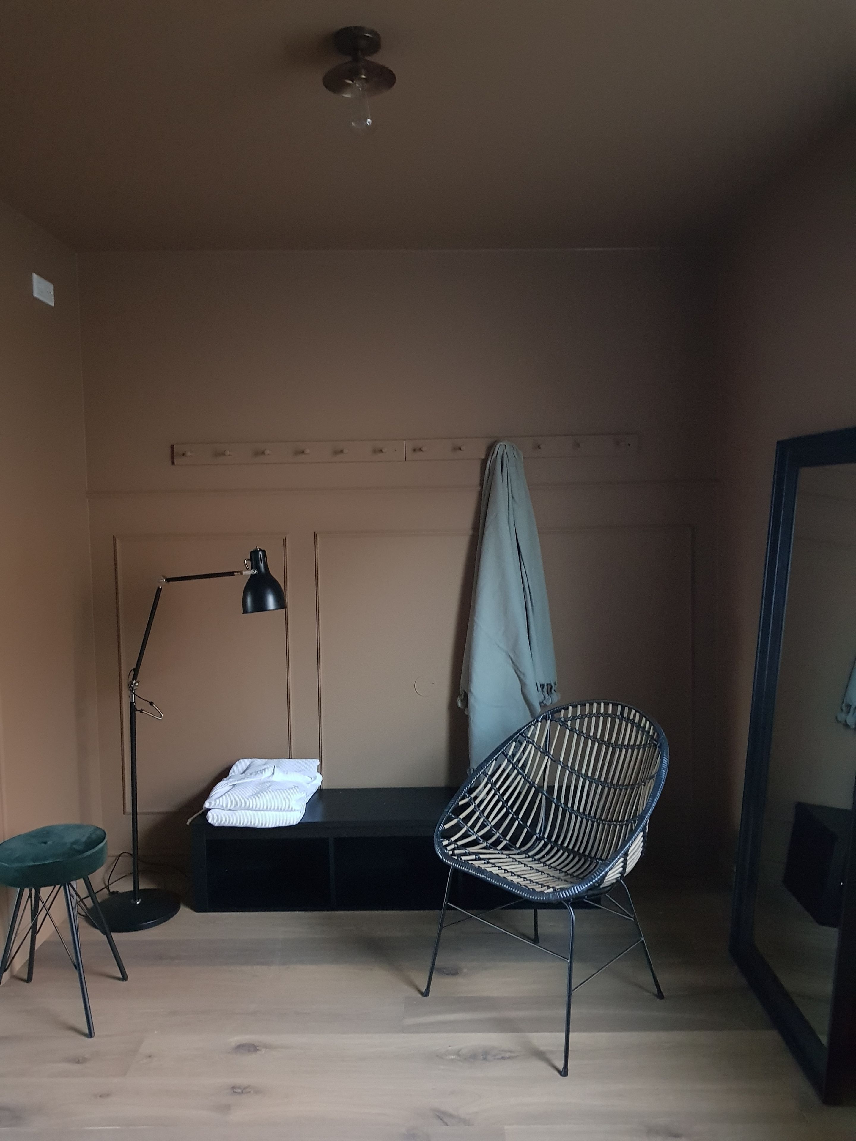 A room with a wooden floor, a black floor lamp, a wicker chair, a bench with a folded towel, and a door with a towel hanging on it.