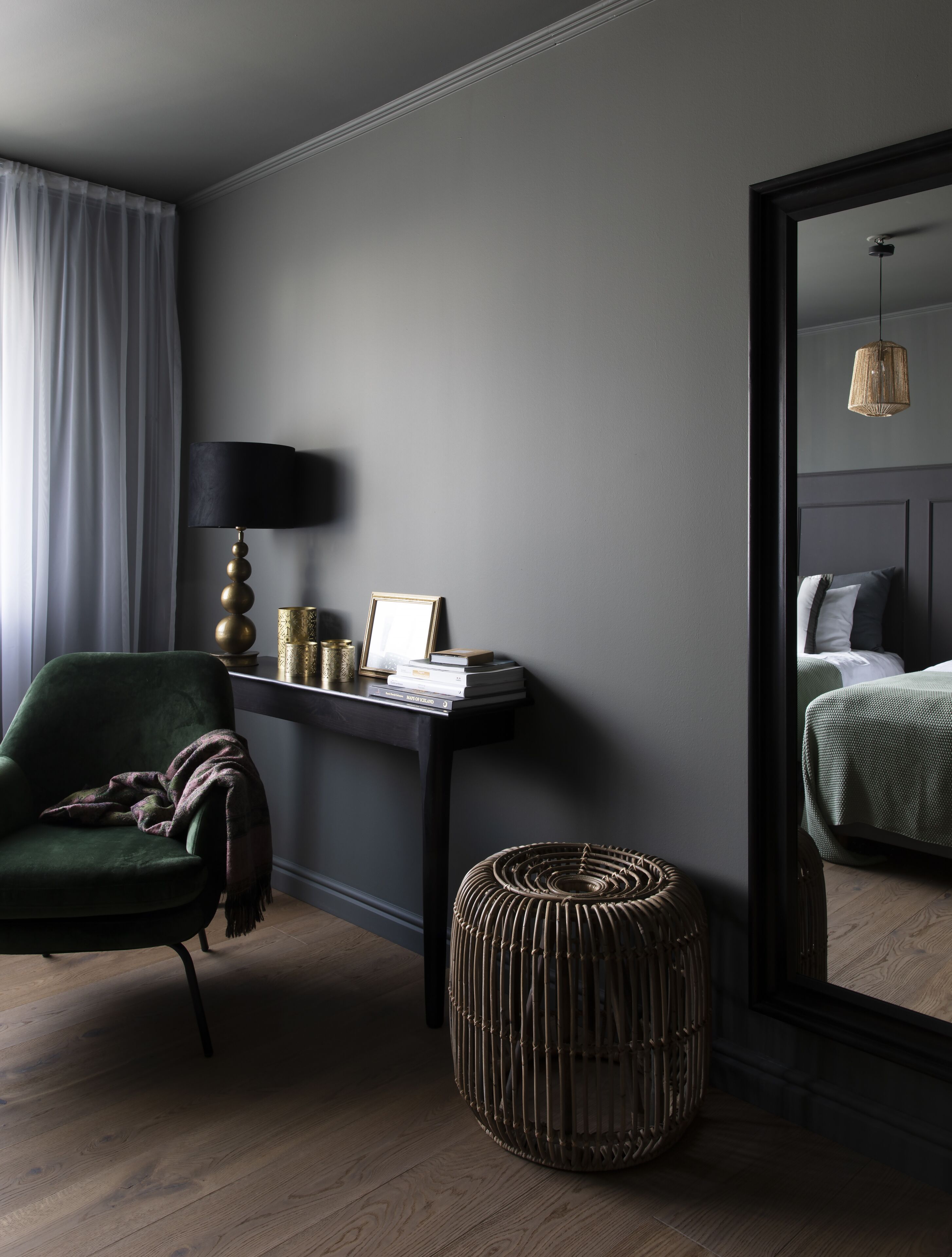 A modern hotel room with a green armchair, a small round ottoman, a desk with a lamp, and a bed with a green bedspread.