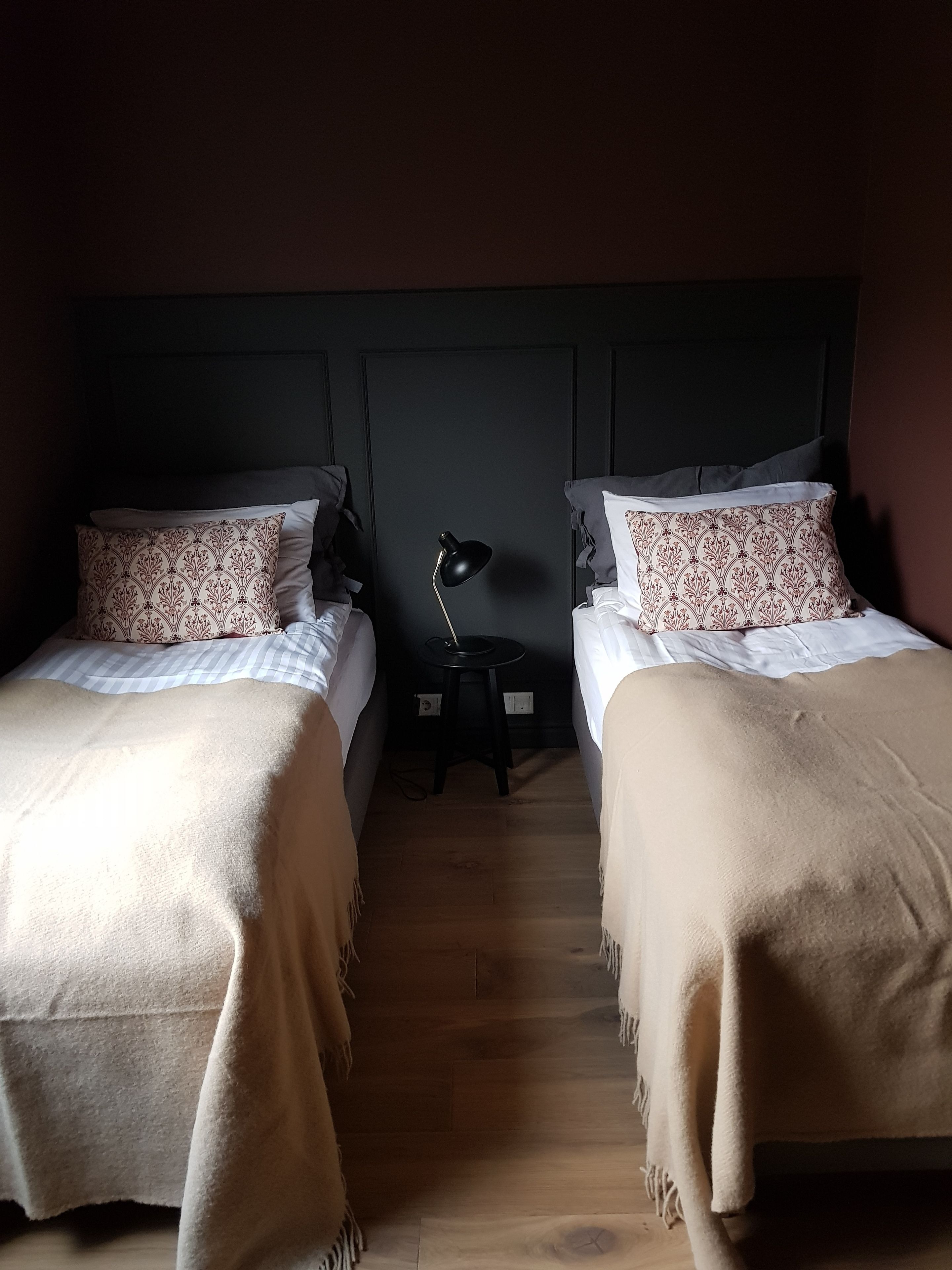 Une chambre avec deux lits, chacun agrémenté d’un coussin à motifs et d’une couverture blanche. Une petite table avec une lampe se trouve entre les deux lits.