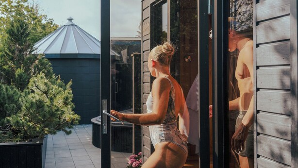 A woman in a swimsuit opening a sliding glass door to a wooden deck with a metal roof structure in the background.
