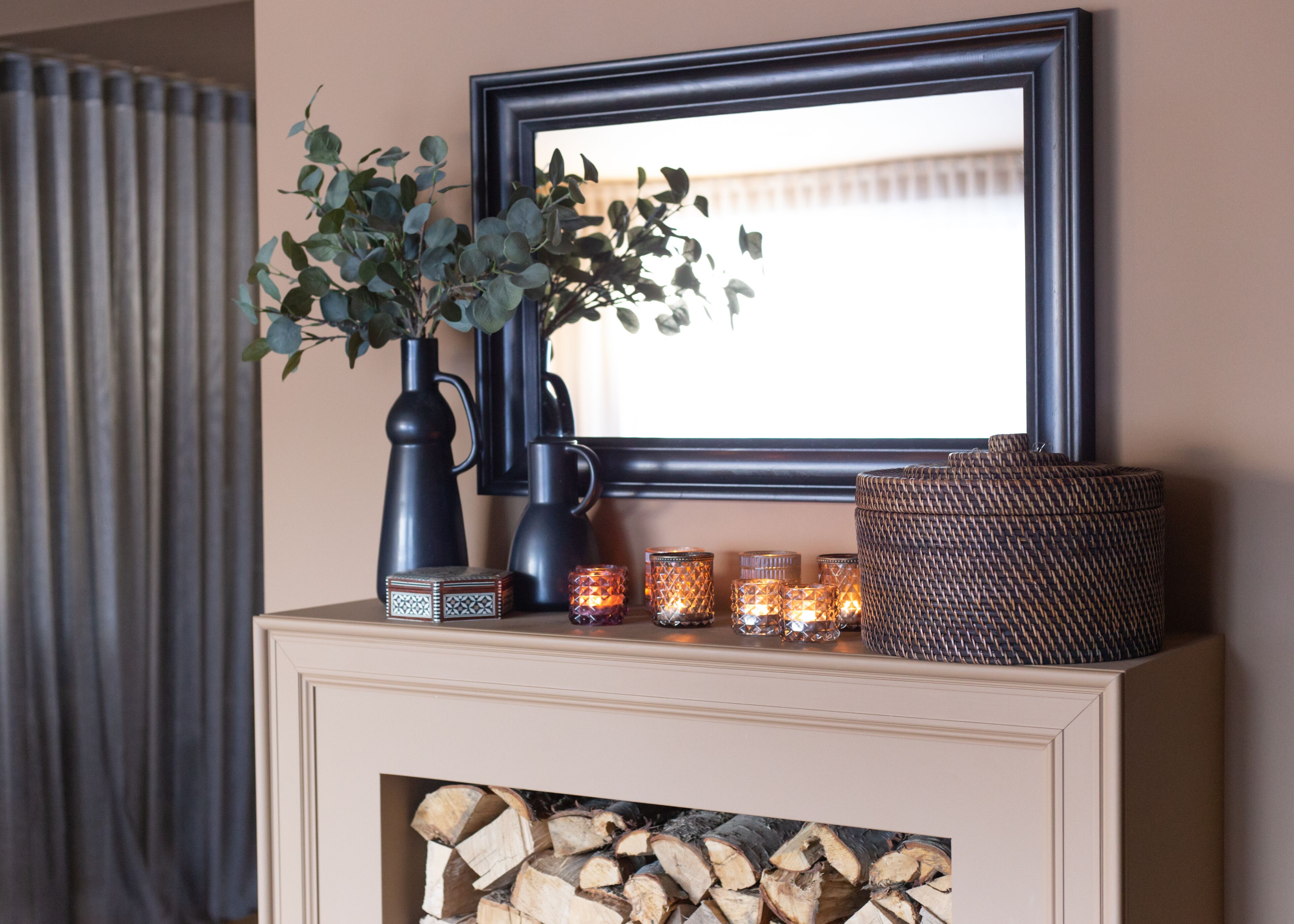 A fireplace mantel with a mirror, a vase with green foliage, a woven basket, and several lit candles.