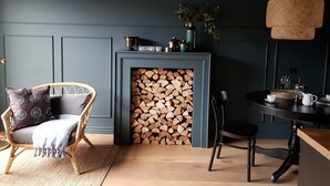 A cozy room with a grey wall, a wicker chair, a small table with a coffee pot, and a fireplace with logs.