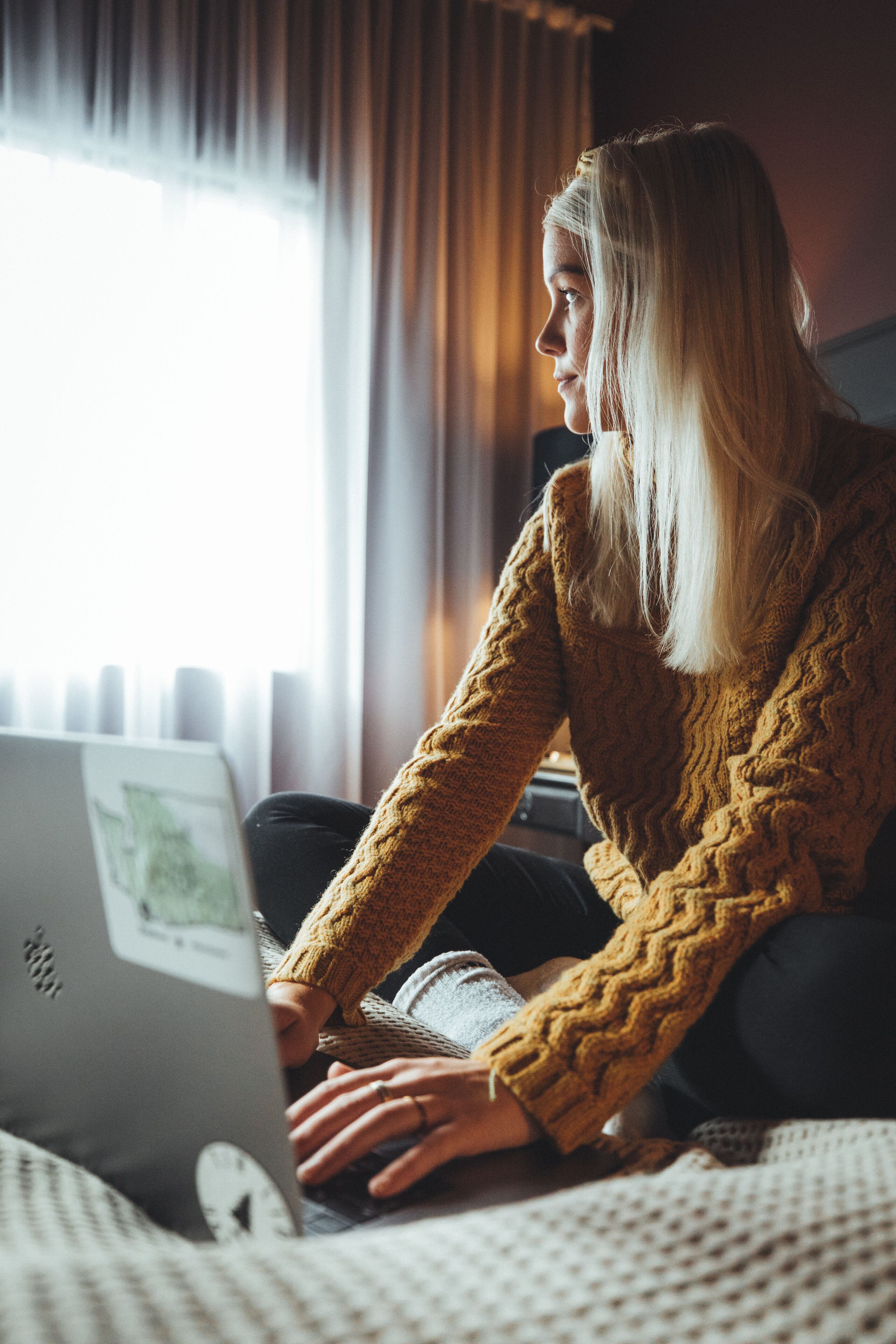 A person using a laptop on a bed with a window in the background.