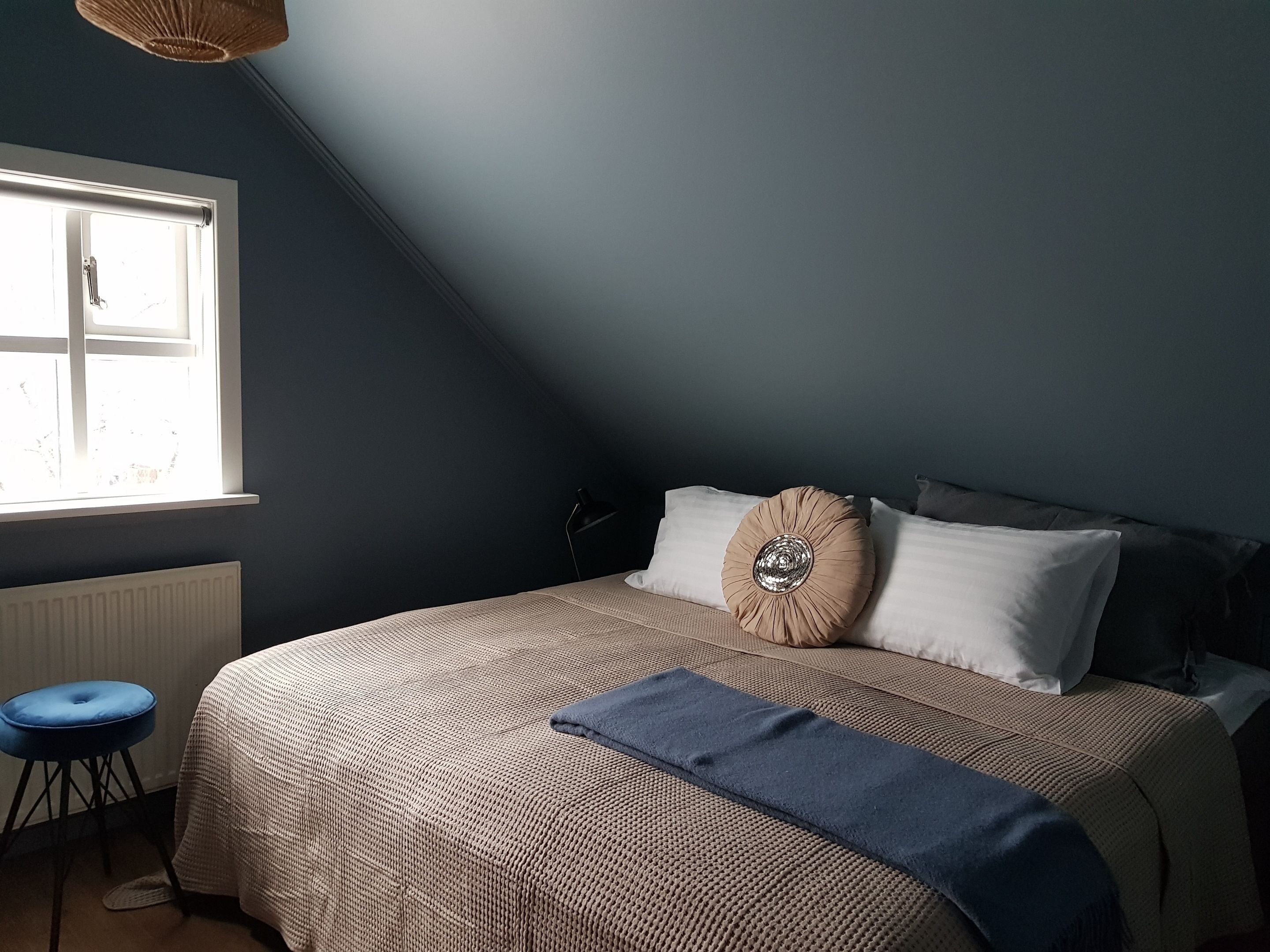 Un dormitorio con una cama grande, una ventana, un radiador y un taburete azul.