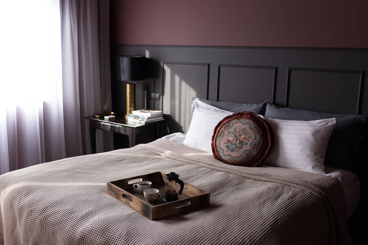 A neatly made bed with a tray of coffee cups and a coffee pot, a bedside table with a lamp, and a window with curtains.