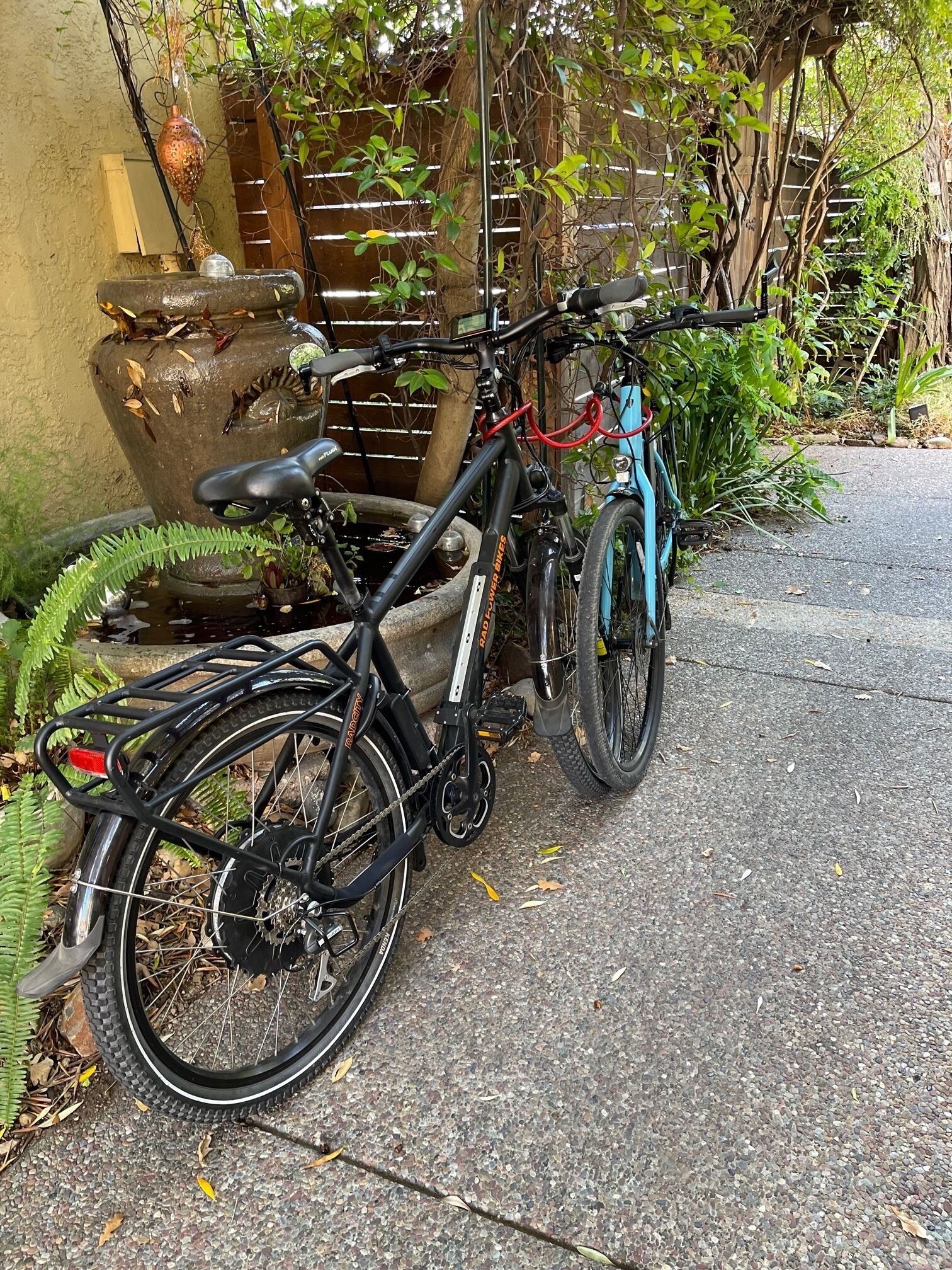 Un vélo est garé à côté d’une fontaine en pierre, dans un jardin verdoyant, délimité par une clôture en bois.