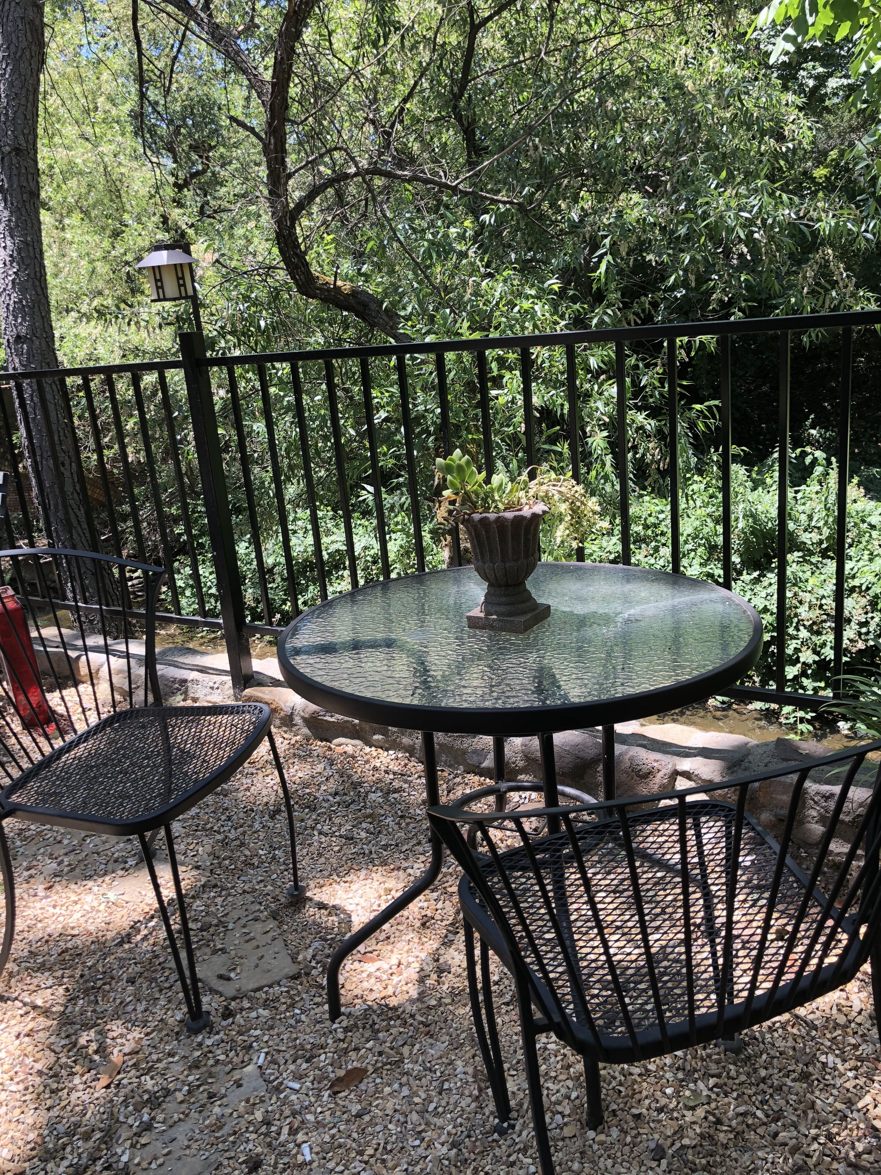 Une table ronde en verre, dotée d’un pied en métal, entourée de chaises en métal, posée sur un sol de gravier, avec une clôture en métal et de la verdure en arrière-plan.
