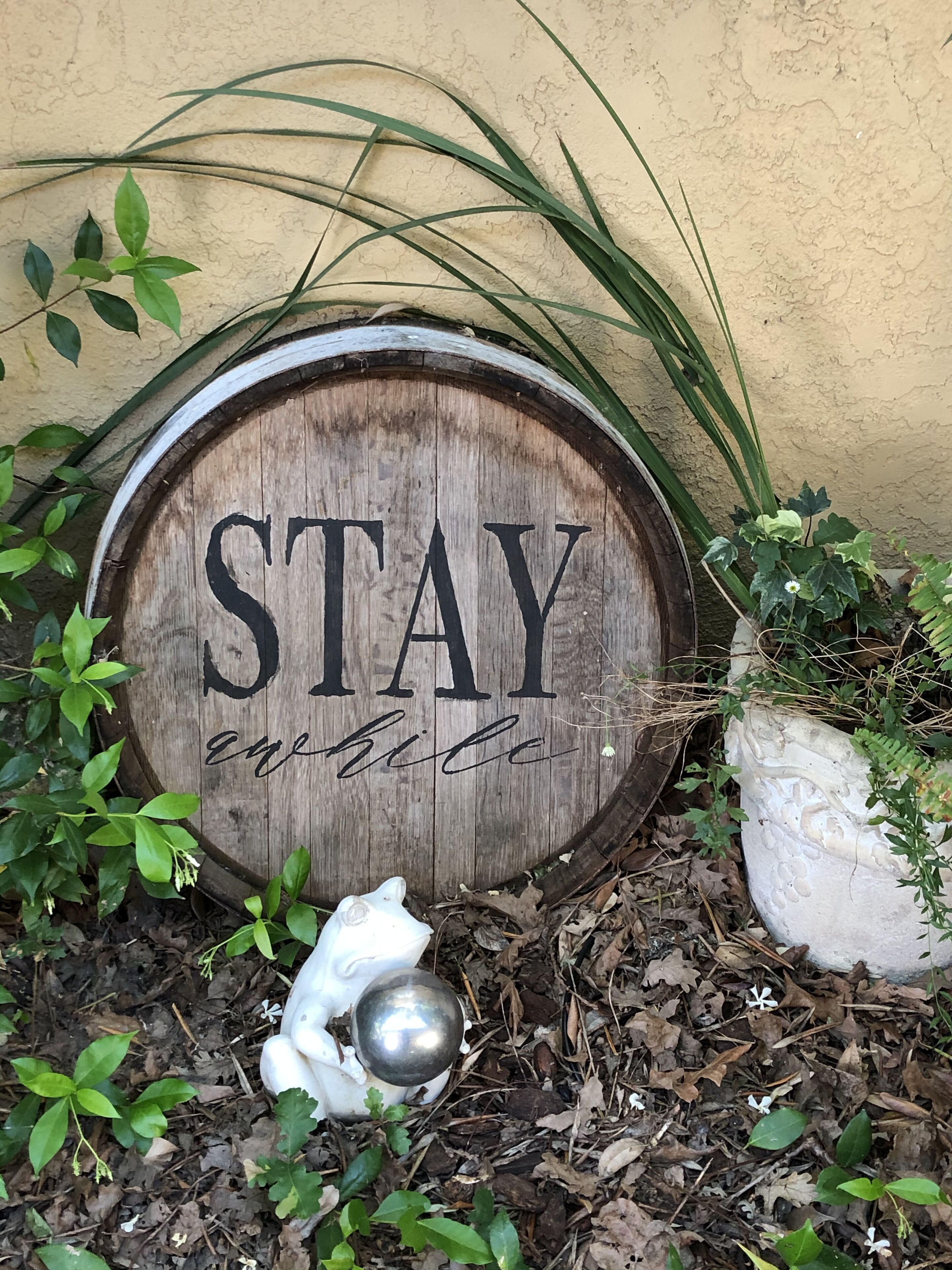 A wooden barrel with the words "STAY" and "enjoy" carved on it, surrounded by greenery and a white decorative object.