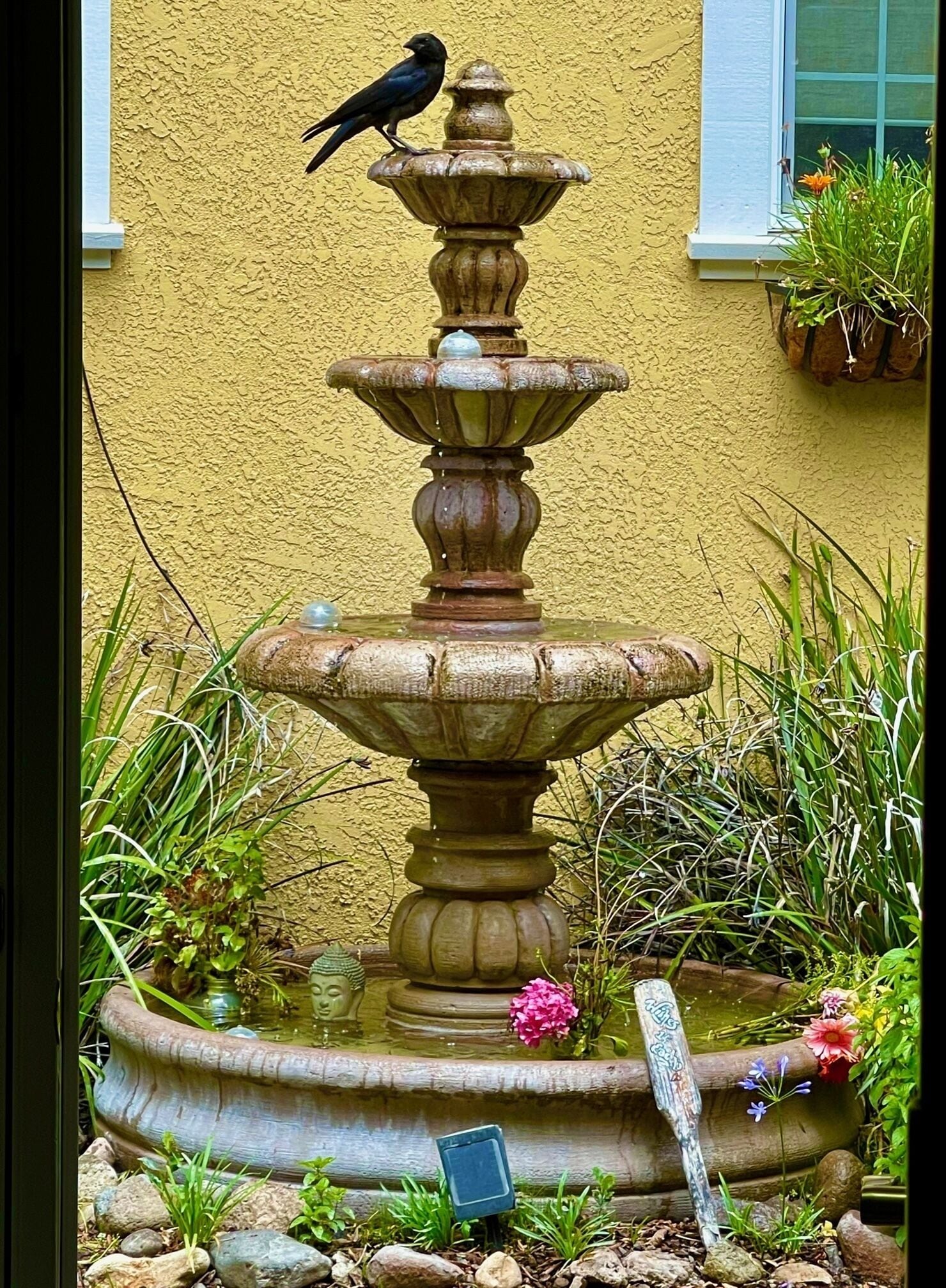 Eine abgestufte Steinfontäne mit einem Vogel auf der obersten Stufe, umgeben von Grünpflanzen und Blumen.