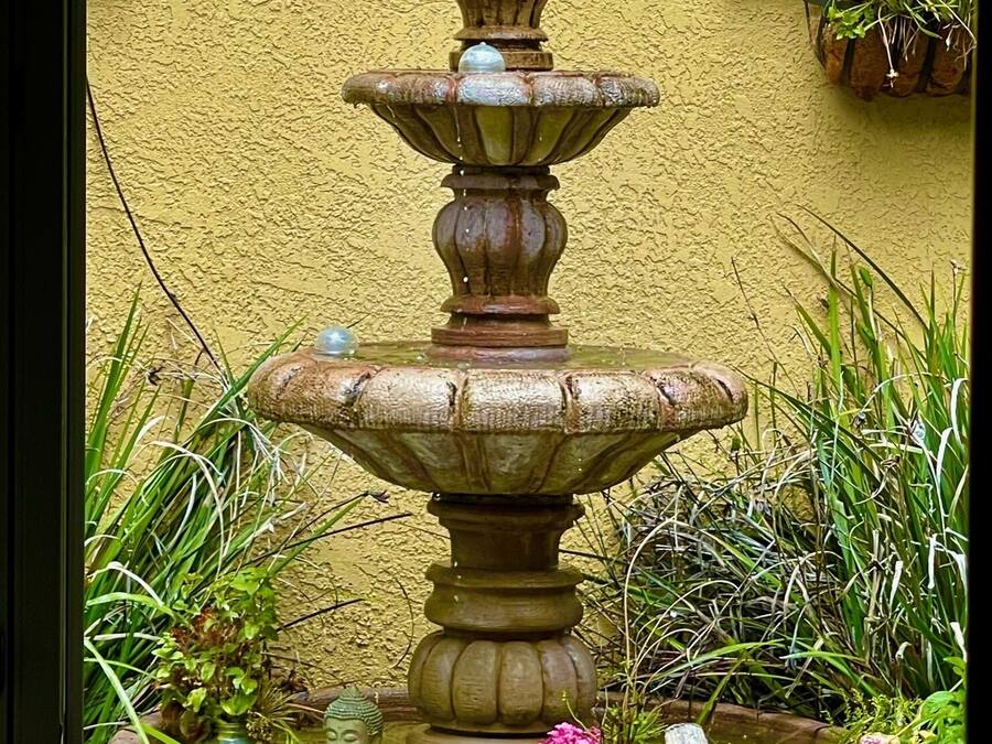 Una fuente de piedra escalonada con un pájaro posado en el nivel superior, rodeada de follaje y flores.