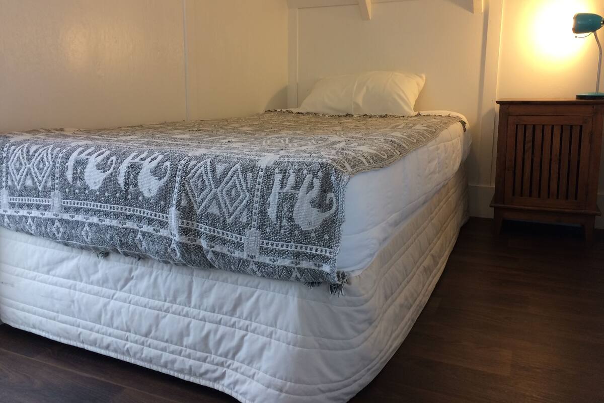 Una cama bien tendida con un edredón estampado, una mesita de noche de madera con una lámpara, y un techo inclinado.