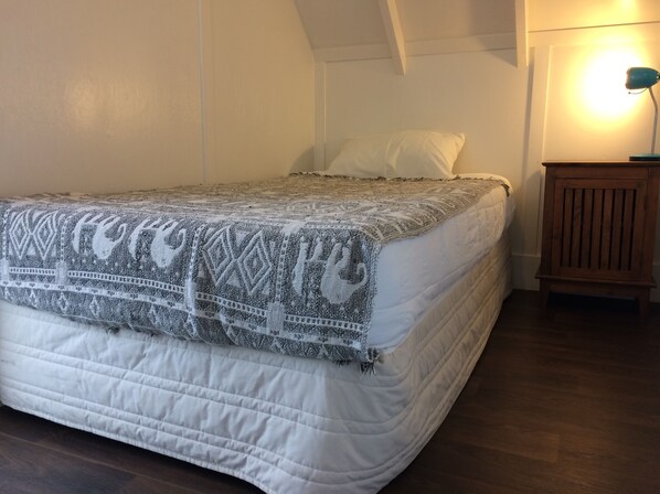 A neatly made bed with a patterned blanket, a wooden nightstand with a lamp, and a sloped ceiling. - Hekerua Lodge Boutique Backpackers (Waiheke Island)