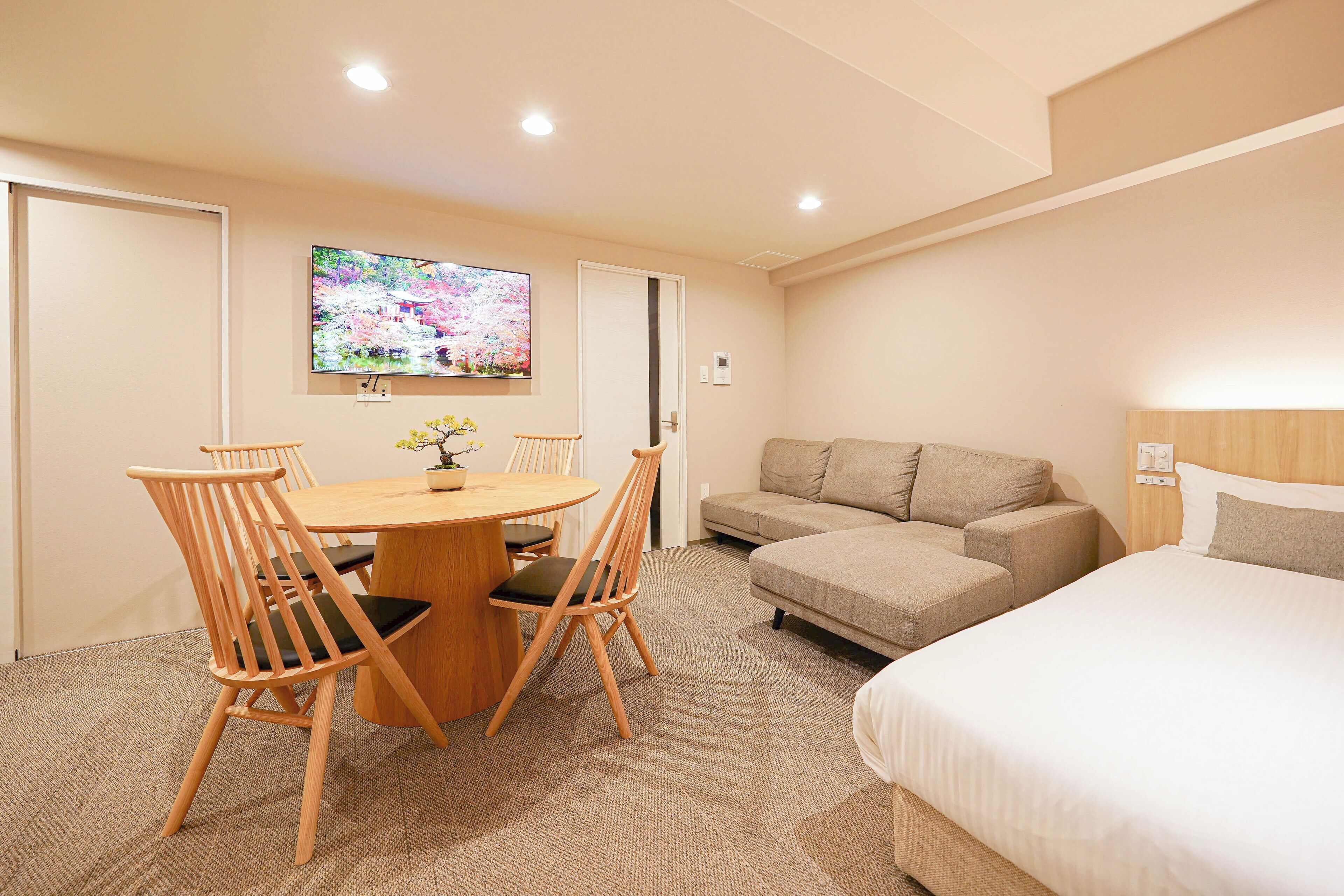 A hotel room with a bed, a sofa, a small table with chairs, a TV on the wall, and a door.