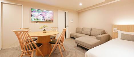 A hotel room with a bed, a sofa, a small table with chairs, a TV on the wall, and a door.