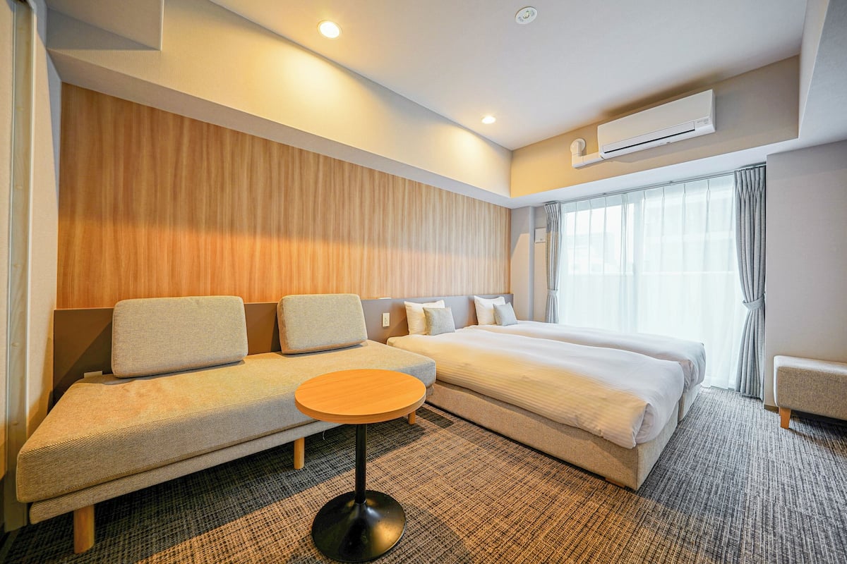 A modern hotel room with a sofa, a bed, a round table, and a large window with curtains.