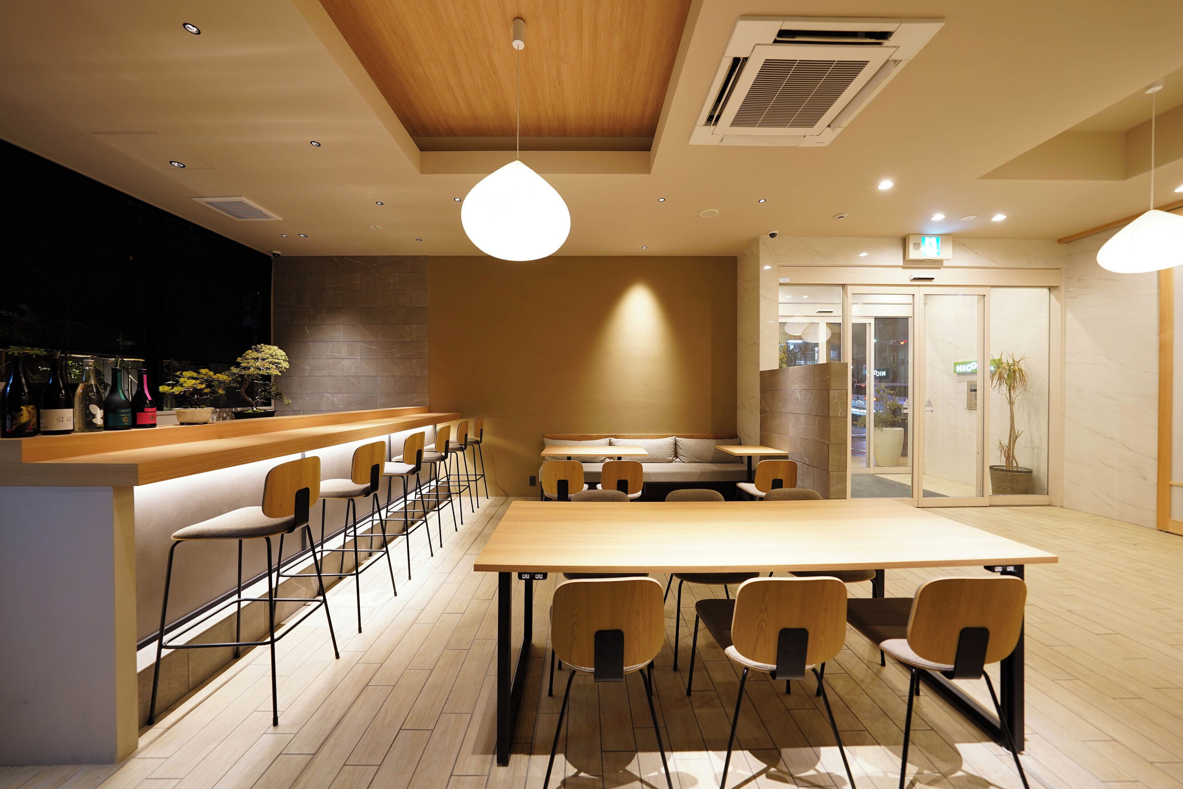 A modern cafe with a wooden bar, multiple seating options including a long table and bar stools, and a casual seating area with a sofa.