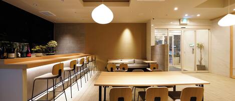 A modern cafe with a wooden bar, multiple seating options including a long table and bar stools, and a casual seating area with a sofa.