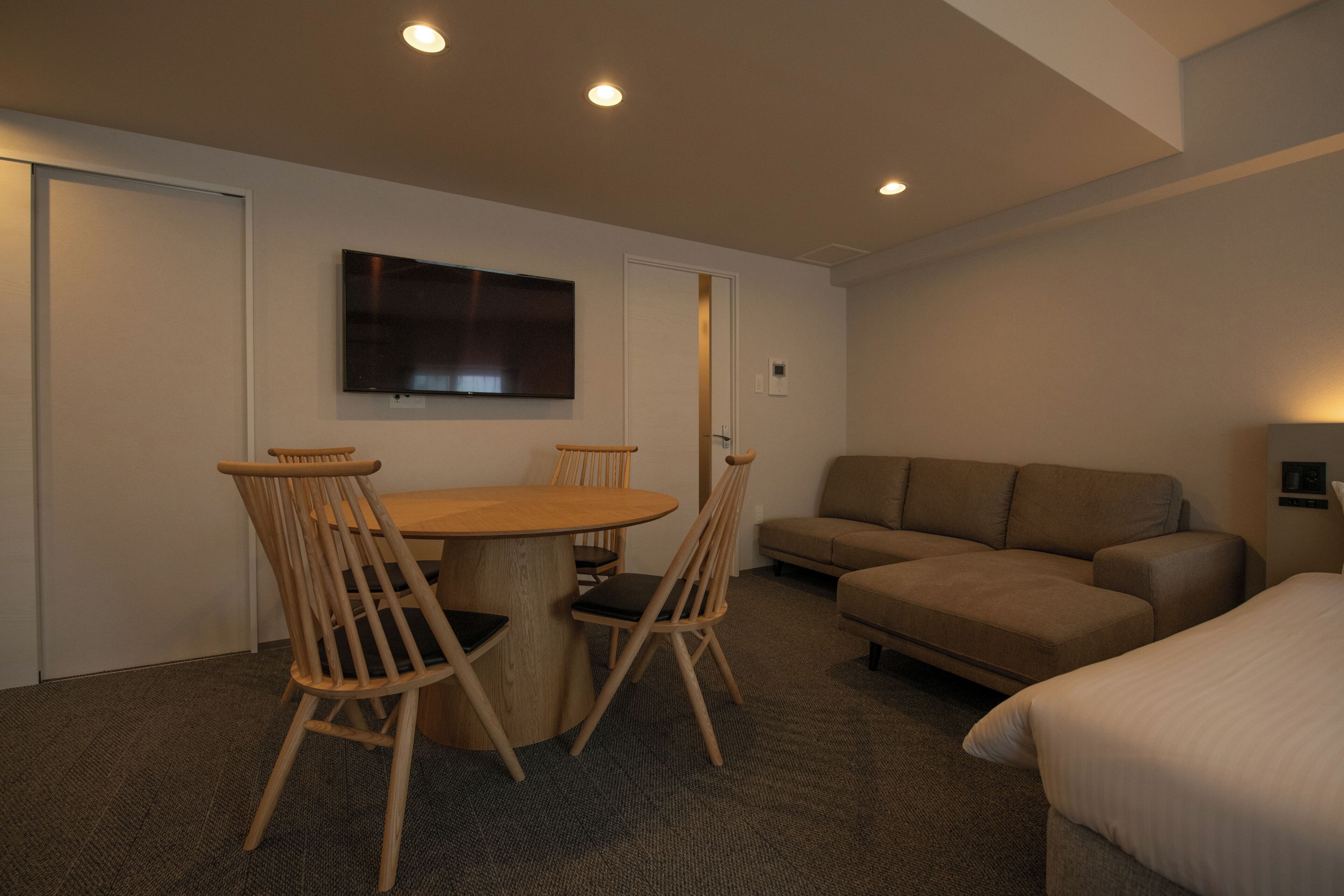 A hotel room with a round wooden table, two wooden chairs, a beige sofa, a flat-screen TV mounted on the wall, and a bed with white bedding.