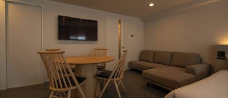 A hotel room with a round wooden table, two wooden chairs, a beige sofa, a flat-screen TV mounted on the wall, and a bed with white bedding.