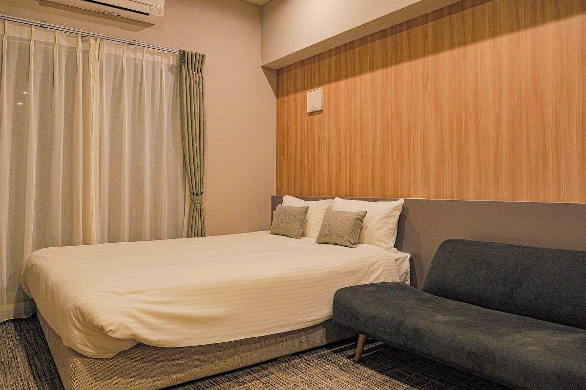 A hotel room with a bed, a grey sofa, a wooden headboard, and an air conditioning unit.