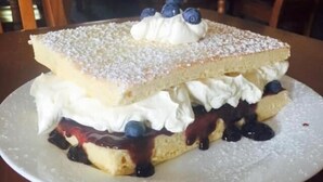 A dessert on a white plate, consisting of a layered cake with whipped cream and blueberries, dusted with powdered sugar.