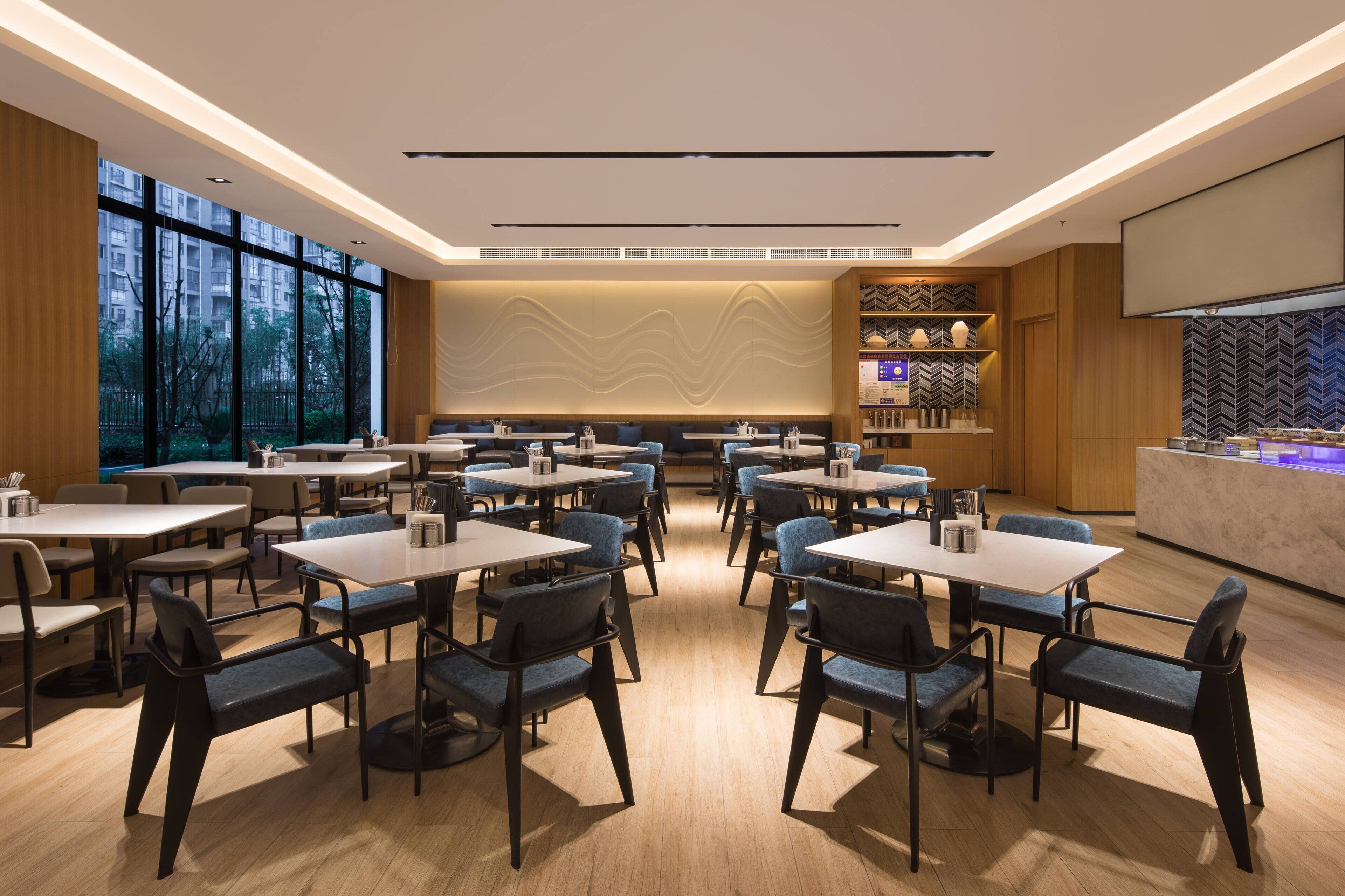 A modern dining area with multiple tables and chairs, large windows, and a kitchen counter.