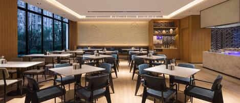 A modern dining area with multiple tables and chairs, large windows, and a kitchen counter.