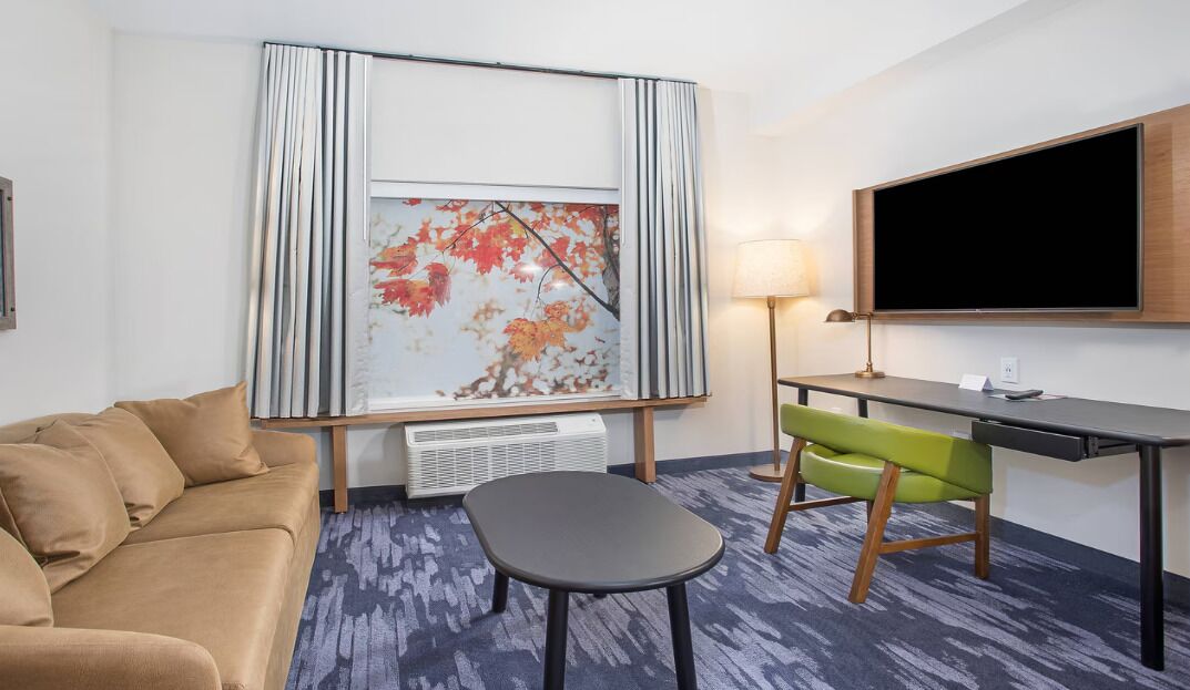 Ein Hotelzimmer mit einer Couch, einem kleinen Tisch, einem Schreibtisch mit Stuhl, einem Flachbildfernseher und einem großen, abstrakten Gemälde an der Wand.