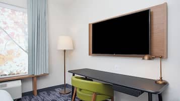 Habitación de hotel con escritorio, una silla verde, televisor de pantalla plana, lámpara y ventana con cortinas.