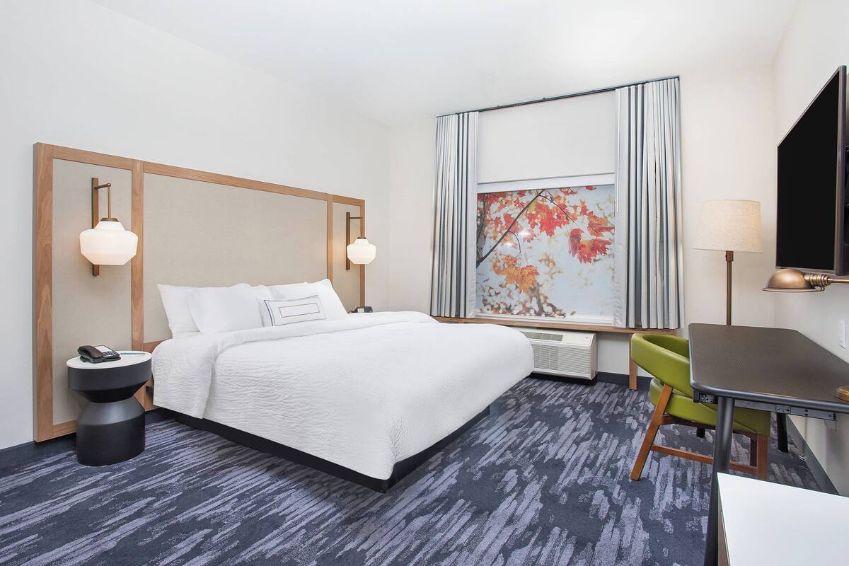 A hotel room with a bed, bedside table, lamp, chair, and a large window with a view.