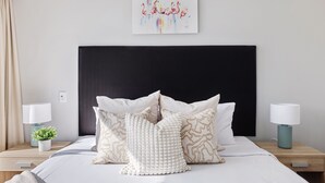 A neatly made bed with white linens and decorative pillows, a black headboard, and bedside tables with lamps and a small plant.